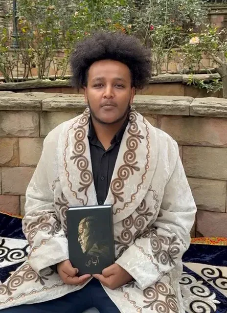 Samson seated outdoors holding a book by Abay while reading his poem.
